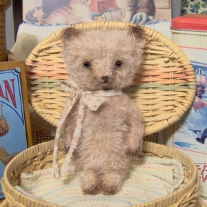 May include: A light brown teddy bear with a white and floral patterned ribbon around its neck, sitting in a wicker chair. The chair is in a wicker basket with a white and pink striped blanket.
