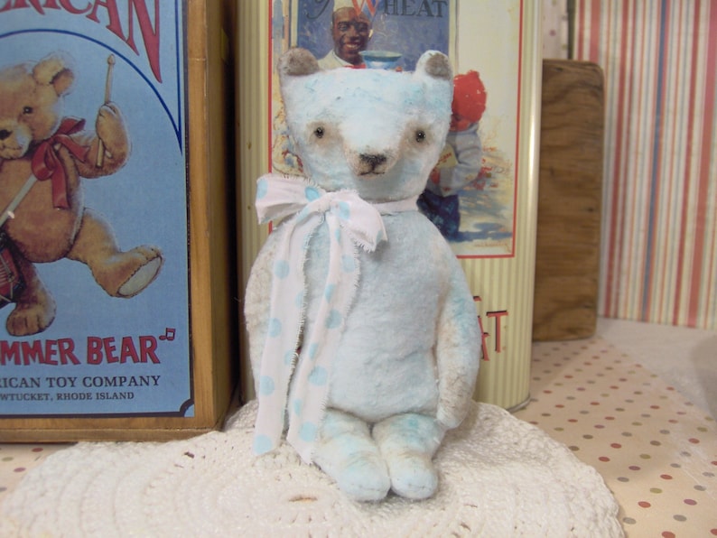 May include: A light blue teddy bear with a white and blue polka dot scarf. The bear has brown eyes and a stitched nose. It is sitting on a white lace doily. The background includes vintage toy packaging.