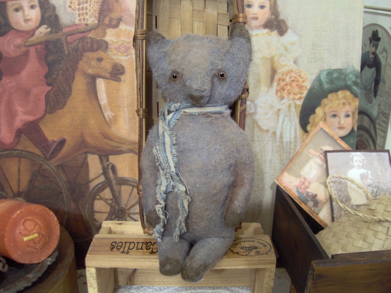 May include: A handmade, gray teddy bear with a blue and white striped scarf. The bear is sitting on a wooden crate labeled "Candles". The background includes vintage-style artwork and a large orange candle.