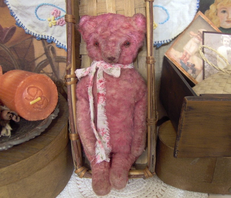 May include: A vintage-style pink teddy bear with a floral ribbon around its neck. The bear is seated in a small, woven chair. The bear has a distressed appearance, suggesting it is an antique or handmade item.