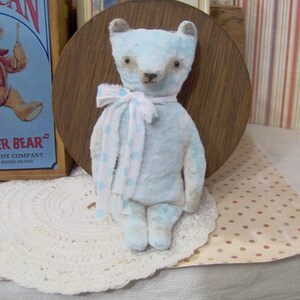 May include: A light blue teddy bear with a white and blue polka dot scarf. The bear has a vintage aesthetic and is sitting on a white lace doily. A vintage "Drummer Bear" sign is in the background.