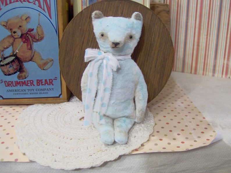 May include: A light blue teddy bear with a white and blue polka dot scarf. The bear has a vintage aesthetic and is sitting on a white lace doily. A vintage "Drummer Bear" sign is in the background.