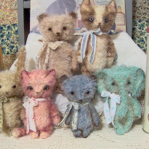 May include: A collection of handmade plush animal toys, including teddy bears, a bunny, and a cat. The toys are in various pastel colors, such as pink, blue, and beige, and are adorned with ribbons. A vintage-style tin is visible in the background.