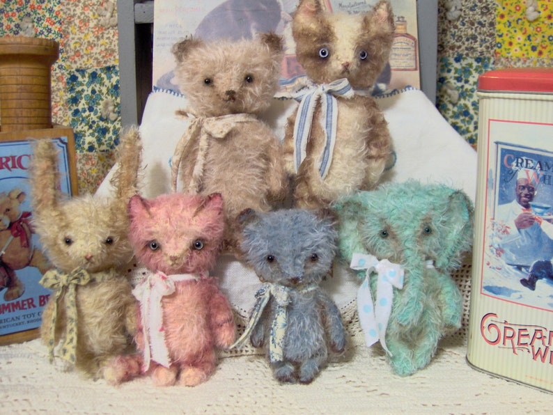 May include: A collection of handmade plush animal toys, including teddy bears, a bunny, and a cat. The toys are in various pastel colors, such as pink, blue, and beige, and are adorned with ribbons. A vintage-style tin is visible in the background.