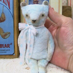 May include: A light blue teddy bear with a white and blue polka dot scarf. The bear has brown eyes and a brown nose. The bear is a vintage toy. The background includes a vintage "Drummer Bear" sign.