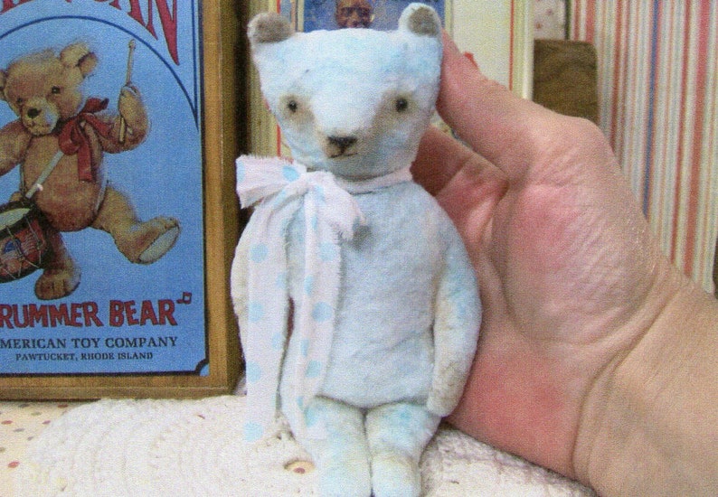 May include: A light blue teddy bear with a white and blue polka dot scarf. The bear has brown eyes and a brown nose. The bear is a vintage toy. The background includes a vintage "Drummer Bear" sign.