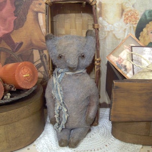 May include: A handmade, vintage-style teddy bear in muted gray tones. The bear has small, dark eyes and a blue and white striped scarf. It stands upright, with a slightly worn appearance, suggesting it's a cherished keepsake. The bear is a charming example of handcrafted toys.
