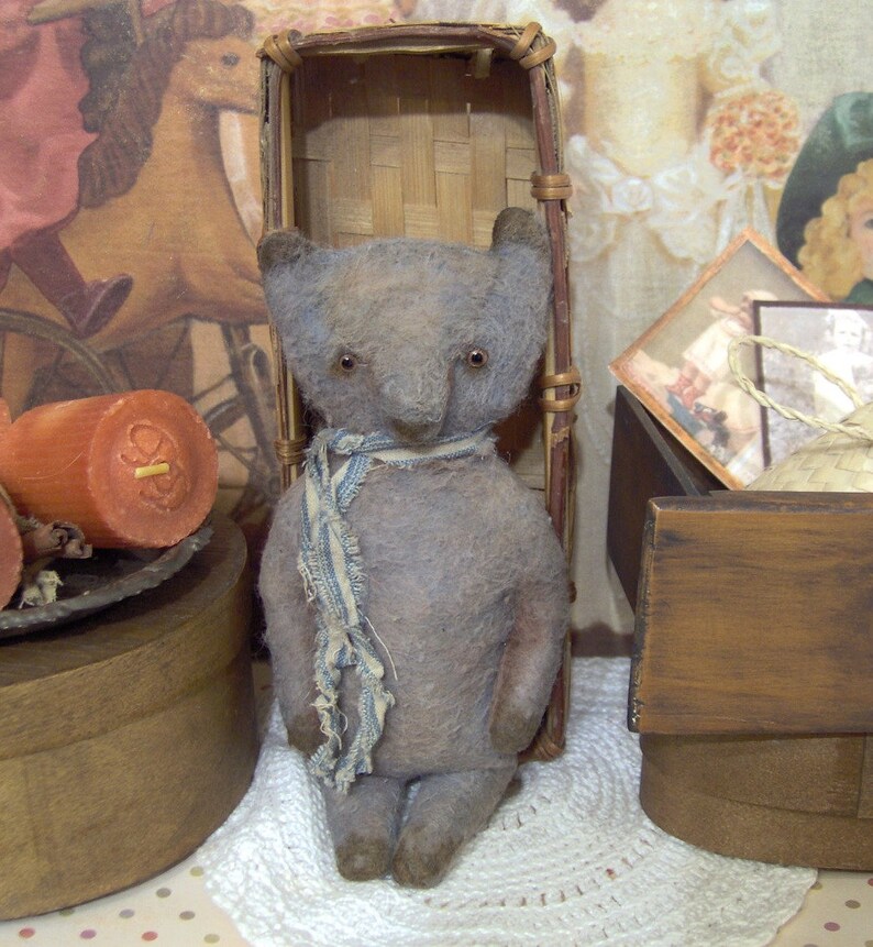 May include: A handmade, vintage-style teddy bear in muted gray tones. The bear has small, dark eyes and a blue and white striped scarf. It stands upright, with a slightly worn appearance, suggesting it's a cherished keepsake. The bear is a charming example of handcrafted toys.