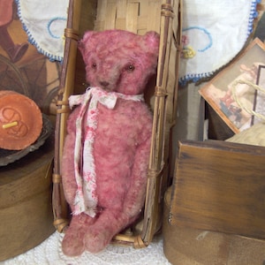 May include: A vintage-style pink teddy bear with a floral ribbon sits in a small, woven basket. The bear has a distressed appearance, with visible seams and a soft texture. The basket is made of light brown woven material. The background includes antique-style decor.