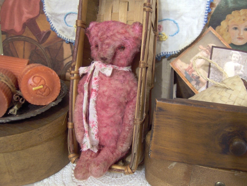 May include: A vintage-style pink teddy bear with a floral ribbon sits in a small, woven basket. The bear has a distressed appearance, with visible seams and a soft texture. The basket is made of light brown woven material. The background includes antique-style decor.