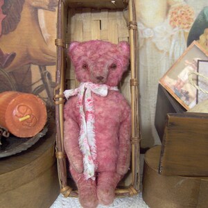 May include: A pink teddy bear with a white floral scarf sits inside a woven basket. The bear has a vintage appearance, with visible stitching and a slightly worn texture. The background includes antique-style decor and candles.