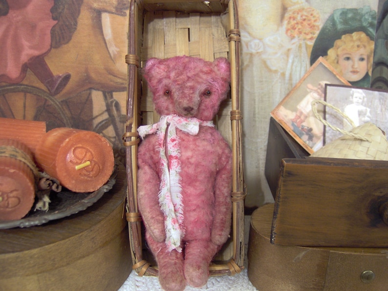 May include: A pink teddy bear with a white floral scarf sits inside a woven basket. The bear has a vintage appearance, with visible stitching and a slightly worn texture. The background includes antique-style decor and candles.
