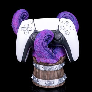 Puede incluir: Un mando blanco y negro de PlayStation 5 es sujetado por tentáculos morados que emergen de un barril de madera. El barril tiene bandas y tachuelas plateadas. El fondo es negro.