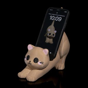 Può includere: Un supporto per telefono a forma di gatto color beige con orecchie rosa e occhi neri. Uno smartphone nero è nel supporto, che mostra un orologio digitale che segna le 10:09. Lo schermo del telefono mostra anche il testo "Passe o dedo para cima para desbloquear" in portoghese.