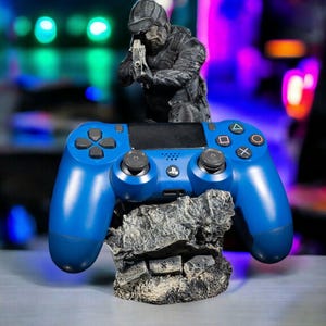 Könnte beinhalten: Ein blauer PlayStation 4-Controller steht auf einem grauen, felsigen Ständer. Auf dem Ständer steht eine schwarze Figur eines Soldaten, der eine Waffe hält.