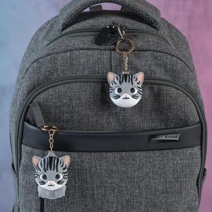 May include: A gray backpack with two cat-shaped keychains attached to the zippers. The keychains are gray and white with black stripes and have pink noses and whiskers.