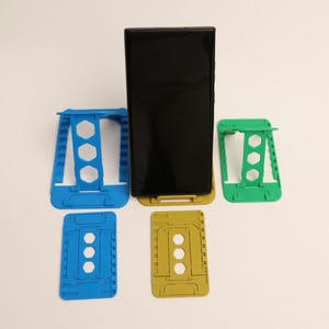 May include: Four phone stands in different colors: blue, green, and yellow. The stands are made of plastic and have a honeycomb design. The stands are designed to hold a smartphone in a vertical position.