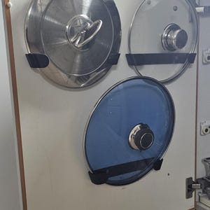 Puede incluir: Una puerta de armario con tres tapas de olla de diferentes estilos. La tapa superior izquierda es de acero inoxidable con un asa de bucle. La tapa superior derecha es de vidrio transparente con un asa de metal. La tapa inferior es de vidrio azul con un asa de metal.