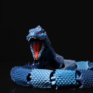 Könnte beinhalten: Eine blaue Spielzeugschlange aus Plastik mit roten Augen und einem segmentierten Körper. Die Schlange ist in einer Pose mit offenem Maul, das ihre Zähne zeigt.