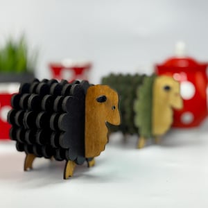 Könnte beinhalten: Holzschaf-Figuren in Schwarz, Grün und Naturholztönen. Die Schafe haben ein geschichtetes Design und stehen auf kleinen Holzbeinen. Eine rote Teekanne mit weißen Punkten ist im Hintergrund.