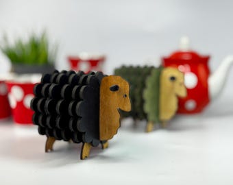 May include: Wooden sheep figurines in black, green, and natural wood tones. The sheep have a layered design and are standing on small wooden legs. A red teapot with white polka dots is in the background.
