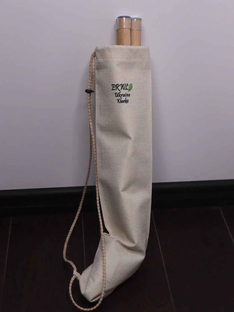 K&ouml;nnte beinhalten: Beige Canvas-Tasche mit Kordelzug, die zwei zylindrische Holzobjekte enth&auml;lt. Auf der Tasche befindet sich ein gr&uuml;nes Logo mit dem Text "ERUL Ukraine Kharkiv". Ein beiges Seil ist befestigt.