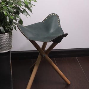 K&ouml;nnte beinhalten: Ein dunkelgr&uuml;ner Leder-Klapphocker mit Messingnieten am Rand des Sitzes. Der Hocker hat ein Holzgestell mit drei Beinen. Die Beine sind f&uuml;r Stabilit&auml;t abgewinkelt. Der Hocker ist f&uuml;r den Transport und den Au&szlig;enbereich konzipiert.