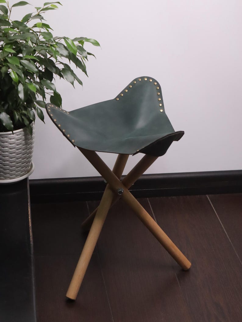 K&ouml;nnte beinhalten: Ein dunkelgr&uuml;ner Leder-Klapphocker mit Messingnieten am Rand des Sitzes. Der Hocker hat ein Holzgestell mit drei Beinen. Die Beine sind f&uuml;r Stabilit&auml;t abgewinkelt. Der Hocker ist f&uuml;r den Transport und den Au&szlig;enbereich konzipiert.