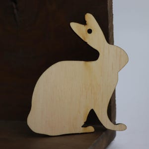 May include: A wooden cutout of a rabbit in a light brown color. The rabbit is facing the right side of the image and has its ears up.