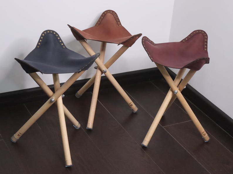 K&ouml;nnte beinhalten: Drei Klapphocker mit Ledersitzen in Marineblau, Braun und Burgunderrot. Die Sitze sind gen&auml;ht und an Holzbeinen befestigt, die sich in der Mitte kreuzen. Die Hocker stehen auf einem dunkelbraunen Holzboden.