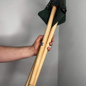 K&ouml;nnte beinhalten: Ein zusammengeklappter Holzstuhl mit dunkelgr&uuml;ner Ledersitzfl&auml;che. Der Hocker hat drei Holzbeine und wird durch ein Metallscharnier zusammengehalten. Der Sitz ist mit kleinen, goldfarbenen Nieten verziert.