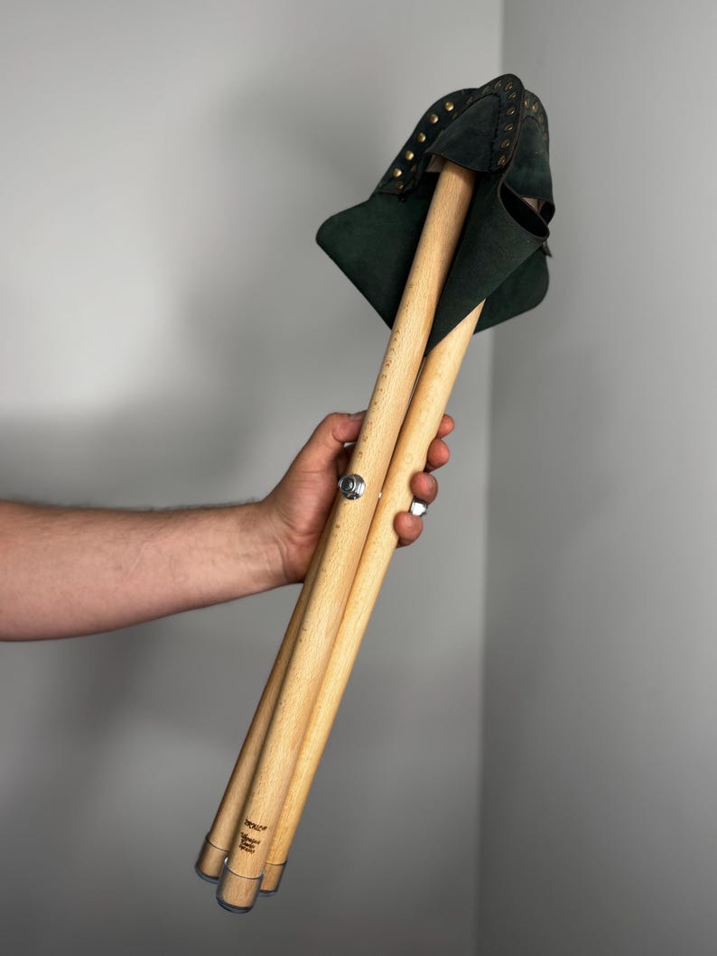 K&ouml;nnte beinhalten: Ein zusammengeklappter Holzstuhl mit dunkelgr&uuml;ner Ledersitzfl&auml;che. Der Hocker hat drei Holzbeine und wird durch ein Metallscharnier zusammengehalten. Der Sitz ist mit kleinen, goldfarbenen Nieten verziert.