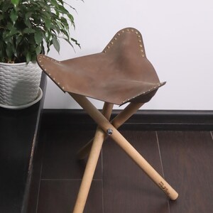 K&ouml;nnte beinhalten: Ein brauner Lederhocker mit einem schmetterlingsf&ouml;rmigen Sitz. Der Sitz ist mit Messingnieten befestigt und ruht auf drei Holzbeinen. Der Hocker ist etwa 46 cm hoch.
