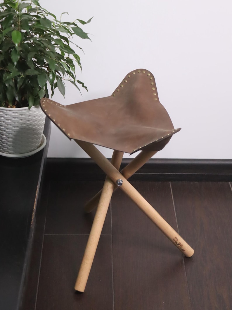 K&ouml;nnte beinhalten: Ein brauner Lederhocker mit einem schmetterlingsf&ouml;rmigen Sitz. Der Sitz ist mit Messingnieten befestigt und ruht auf drei Holzbeinen. Der Hocker ist etwa 46 cm hoch.