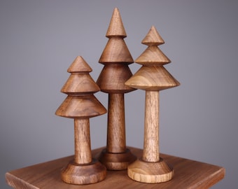 Puede incluir: Tres figuras de árboles de Navidad de madera de diferentes alturas. Los árboles son de madera marrón claro y tienen un diseño en capas. Se exhiben sobre una superficie de madera sobre un fondo gris.