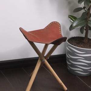K&ouml;nnte beinhalten: Ein kleiner, zusammenklappbarer Hocker mit braunem Ledersitz und Holzbeinen. Der Sitz ist mit kleinen, goldfarbenen Nieten befestigt. Die Beine sind aus hellem Holz, und der Hocker steht vor einer wei&szlig;en Wand und einem dunklen Holzboden.