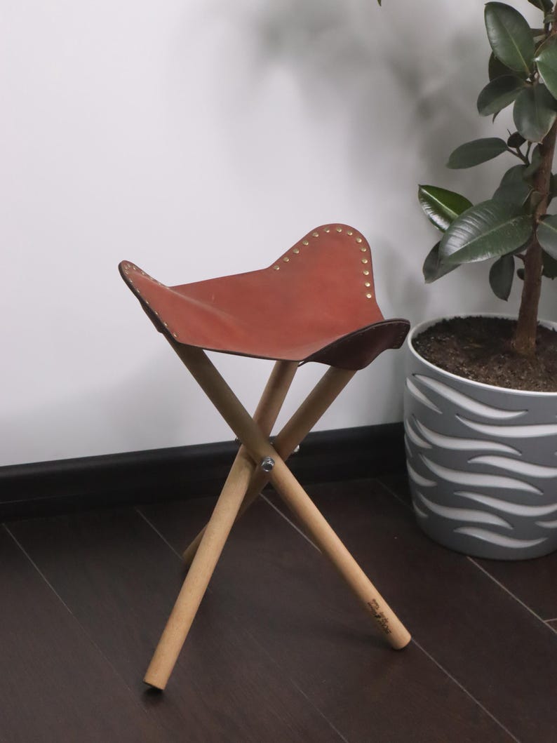 K&ouml;nnte beinhalten: Ein kleiner, zusammenklappbarer Hocker mit braunem Ledersitz und Holzbeinen. Der Sitz ist mit kleinen, goldfarbenen Nieten befestigt. Die Beine sind aus hellem Holz, und der Hocker steht vor einer wei&szlig;en Wand und einem dunklen Holzboden.