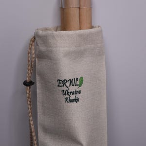 K&ouml;nnte beinhalten: Ein beigefarbener Kordelzugbeutel mit dem Text "ERWL Ukraine Kharkiv" in Gr&uuml;n. Die Tasche enth&auml;lt zwei Holzd&uuml;bel mit transparenten Kunststoffkappen. Der Kordelzug ist hellbraun.