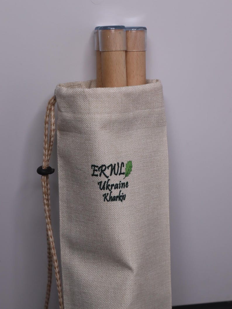 K&ouml;nnte beinhalten: Ein beigefarbener Kordelzugbeutel mit dem Text "ERWL Ukraine Kharkiv" in Gr&uuml;n. Die Tasche enth&auml;lt zwei Holzd&uuml;bel mit transparenten Kunststoffkappen. Der Kordelzug ist hellbraun.
