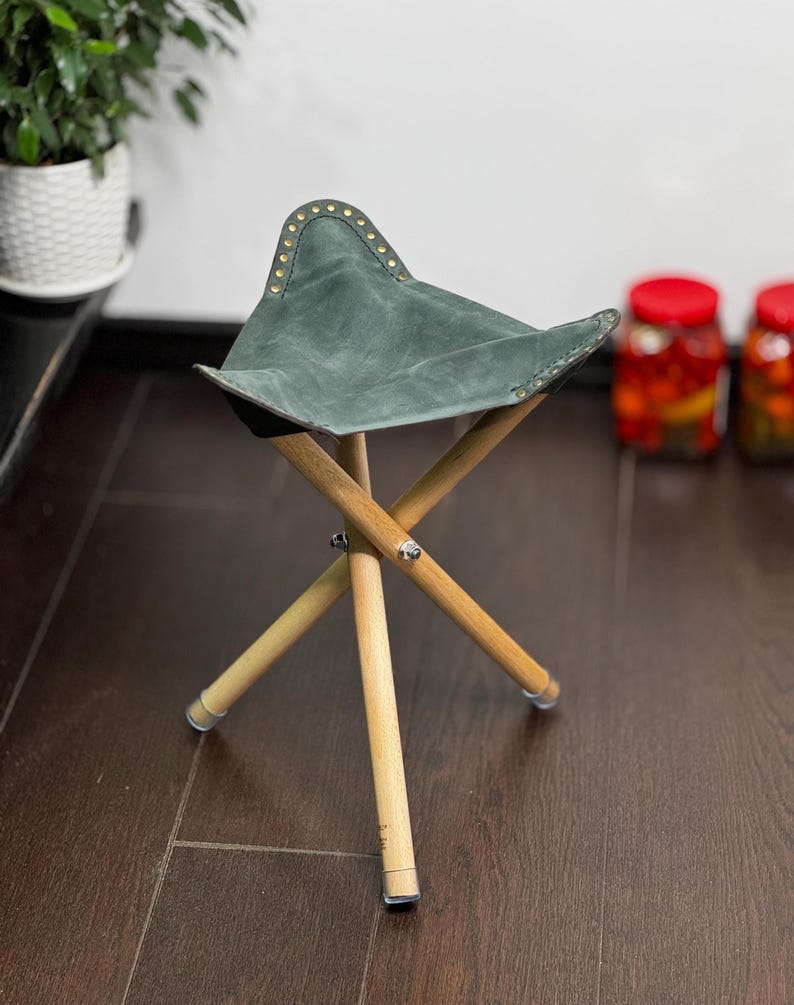 K&ouml;nnte beinhalten: Ein kleiner, zusammenklappbarer Hocker mit einem Sitz aus teal-farbenem Wildleder und Holzbeinen. Der dreieckige Sitz ist mit Messingnieten am Rand verziert. Die hellen Holzbeine sind klappbar.