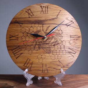 May include: A wooden clock with Roman numeral markings and a bee design on a honeycomb background. The clock is on a clear plastic stand.