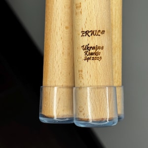 K&ouml;nnte beinhalten: Nahaufnahme von mehreren Holzd&uuml;beln mit transparenten Kunststoff-Endkappen. Der Text "ERWUL" und "Ukraine Kharkiv Sept 2023" ist in das Holz graviert. Die D&uuml;bel sind hellbraun.