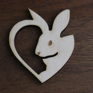 May include: A wooden heart-shaped ornament with a cutout design of a rabbit. The rabbit is facing left and has a simple, stylized design.
