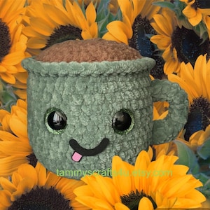 May include: A green crocheted mug with a brown top, smiling with googly eyes and a pink tongue sticking out. The mug is surrounded by yellow sunflowers.