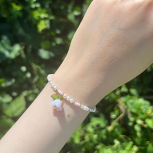 Puede incluir: Una delicada pulsera con perlas blancas de imitación, cuentas doradas y detalles rosas. Un pequeño dije de flor azul claro con hojas verdes cuelga de la pulsera. La pulsera se lleva en una muñeca sobre un fondo de follaje verde.