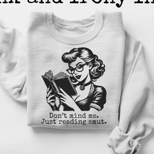 Può includere: Una felpa bianca con un'illustrazione in bianco e nero di una donna che legge un libro. Il testo "Don't mind me. Just reading smut." è stampato sotto l'illustrazione.