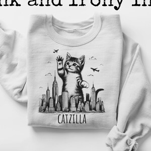 May include: A white sweatshirt with a black and white illustration of a cat standing on a city skyline. The text "CATZILLA" is printed below the illustration.