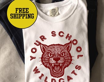 May include: White crew neck t-shirt with a maroon graphic of a snarling wildcat and the words "YOUR SCHOOL WILDCATS" in a circular design. The shirt is folded on top of a pair of blue jeans.