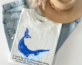 May include: White t-shirt with a blue whale illustration and the quote "I love to sail forbidden seas, and land on barbarous coasts. Herman Melville: Moby-Dick." The shirt is paired with light blue jeans and a tan hat.