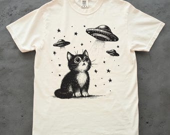 May include: A white t-shirt with a black and white illustration of a cat looking up at a UFO. The UFO is surrounded by stars. The text "Cosmic Cat" is printed on the shirt.